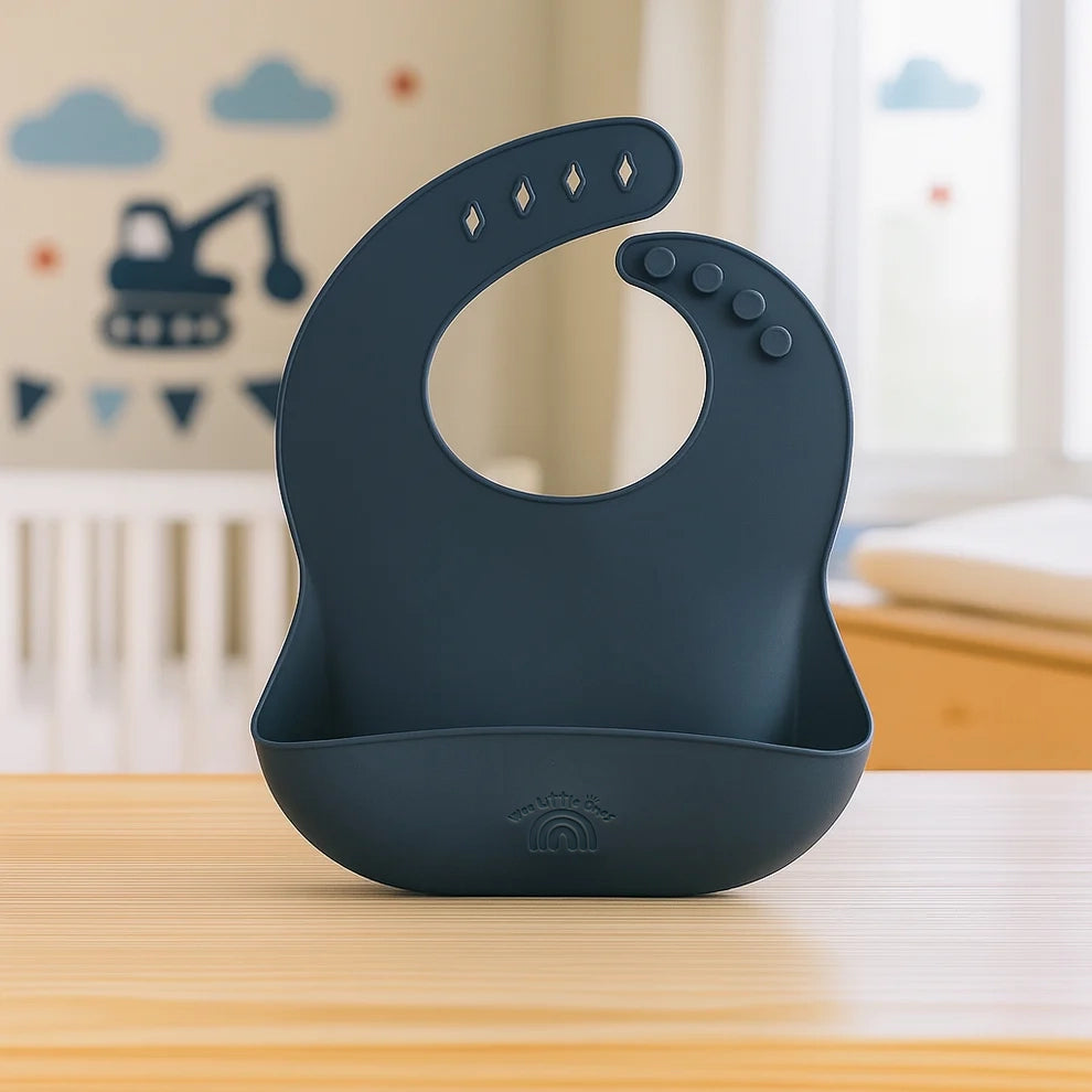 Navy blue silicone bib with adjustable button neck and food catcher pouch on wooden surface.