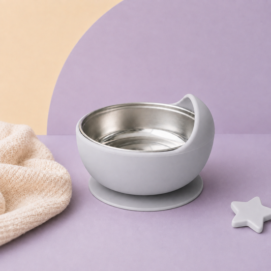 A baby feeding bowl with a matte gray silicone exterior, a stainless steel interior, and a suction base, styled on a beige surface with a cream textured cloth and a wooden star teether.