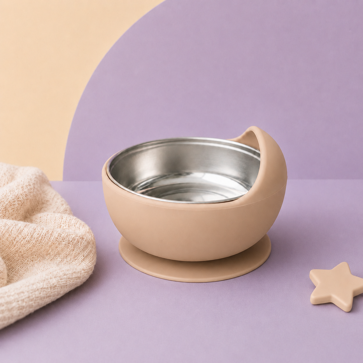 A beige baby feeding bowl with a stainless steel interior and suction base, styled on a pastel background with a white cloth and wooden teething toys.