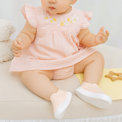 Baby wearing light pink soft-soled breathable toddler shoes with white soles.
