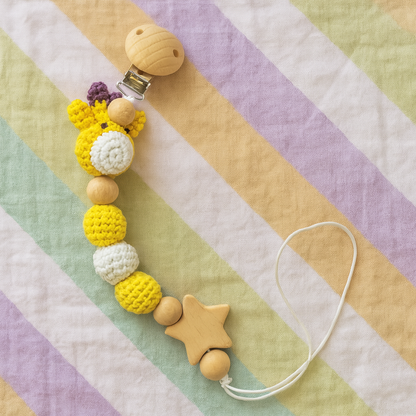 Crochet giraffe pacifier clip with yellow and white yarn beads, wooden star and clip on pastel striped fabric background.