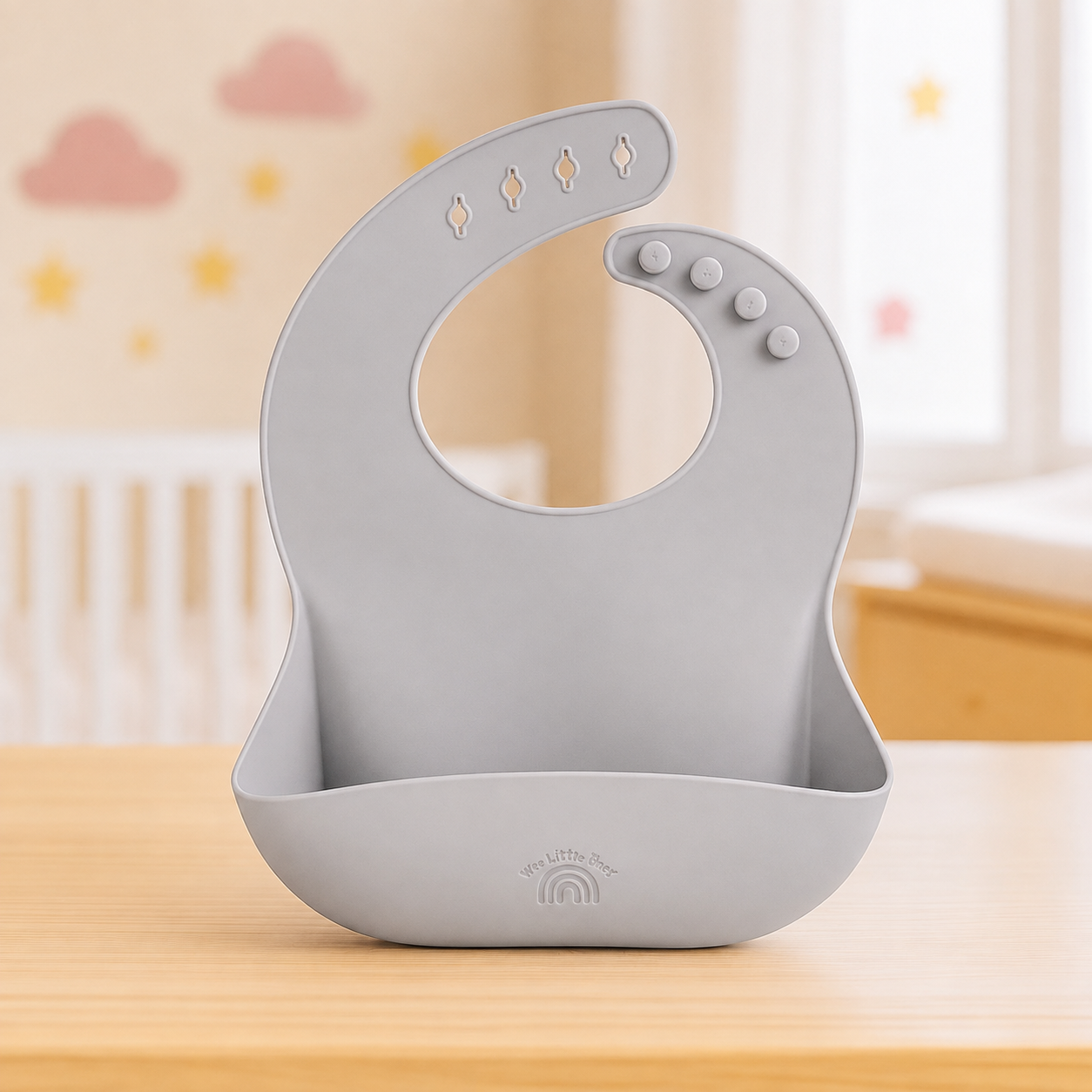 A light gray silicone baby bib and mouth tray set with a curved neck opening, decorative cutouts, and a suction cup base, displayed on a wooden table in a softly blurred nursery setting.