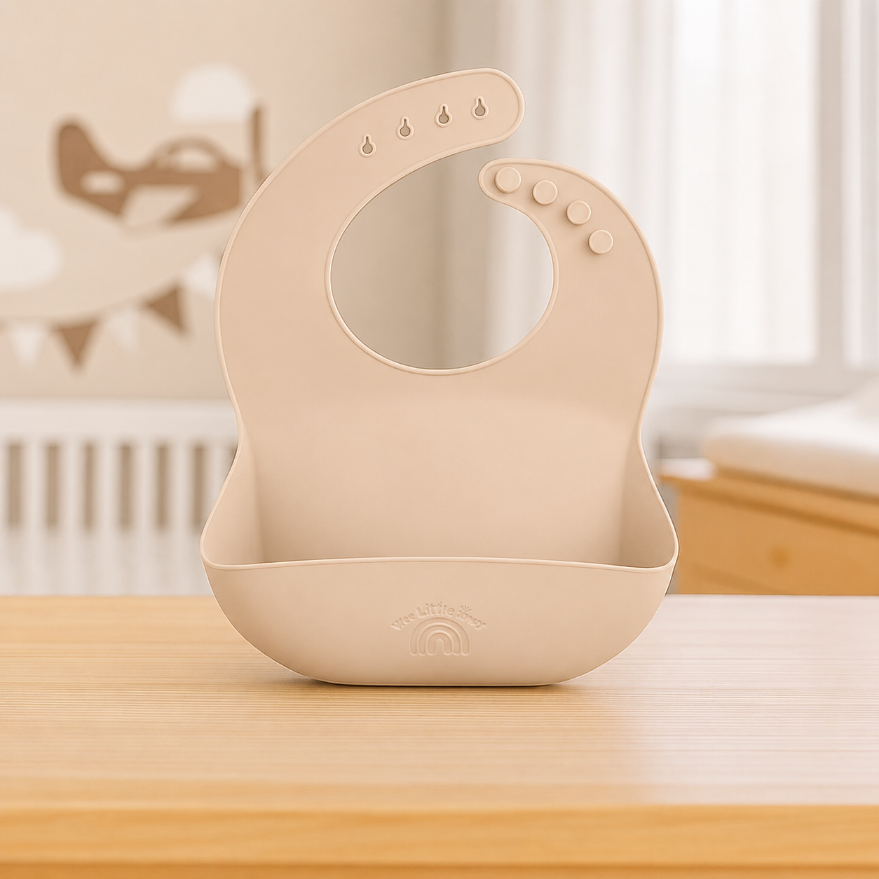 A beige silicone baby bib with a matching bowl, placed on a light wooden table in a softly blurred nursery setting.