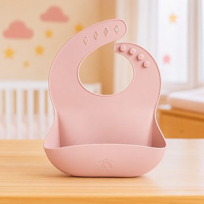 A pink silicone baby bib and mouth guard set with suction cups, shown on a wooden table in a softly blurred nursery setting.
