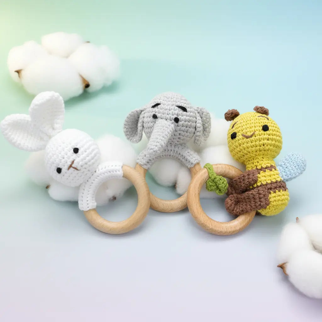 Crocheted white bunny, gray elephant, and yellow bee baby teething rings with wooden handles on light blue background.