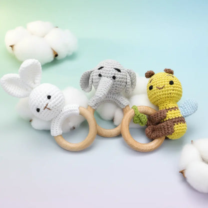 Crocheted white bunny, gray elephant, and yellow bee baby teething rings with wooden handles on light blue background.