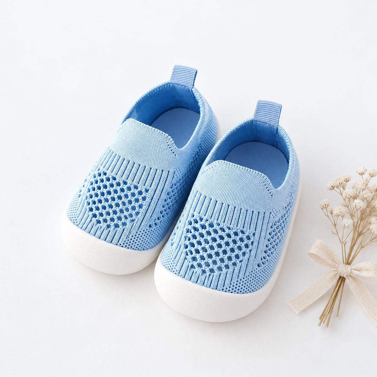 A pair of light blue knit slip-on baby shoes with white soles, arranged on a clean white background next to a small decorative dried flower sprig tied with a cream ribbon.