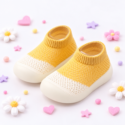 A pair of baby slip-on knit socks with yellow knit uppers, white mesh toe panels, and white soles, styled on a white background with pastel decorative flowers and hearts.