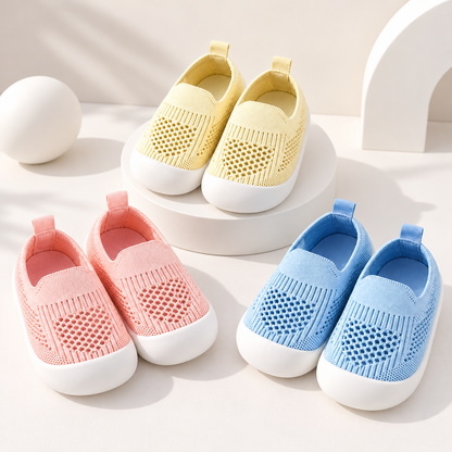 Four slip-on mesh baby shoes in pastel yellow, pink, and light blue, arranged on white display blocks and the floor against a clean white studio background.