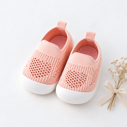 A pair of baby slip-on shoes in a soft peach-pink color with white rubber soles, styled on a clean white background next to a small decorative dried flower sprig.