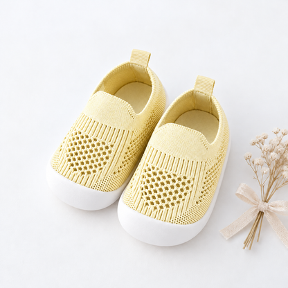 A pair of baby slip-on shoes in pale yellow knit fabric with white soles, arranged on a white background next to a small decorative dried flower sprig.