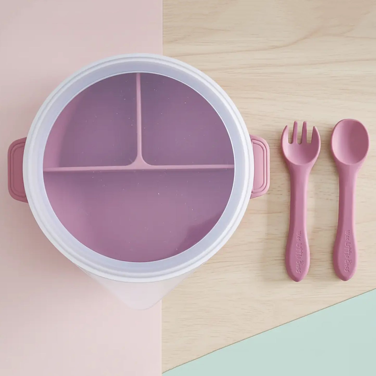 Round dark pink silicone divided plate with lid, matching spoon and fork set on wood and pastel background.
