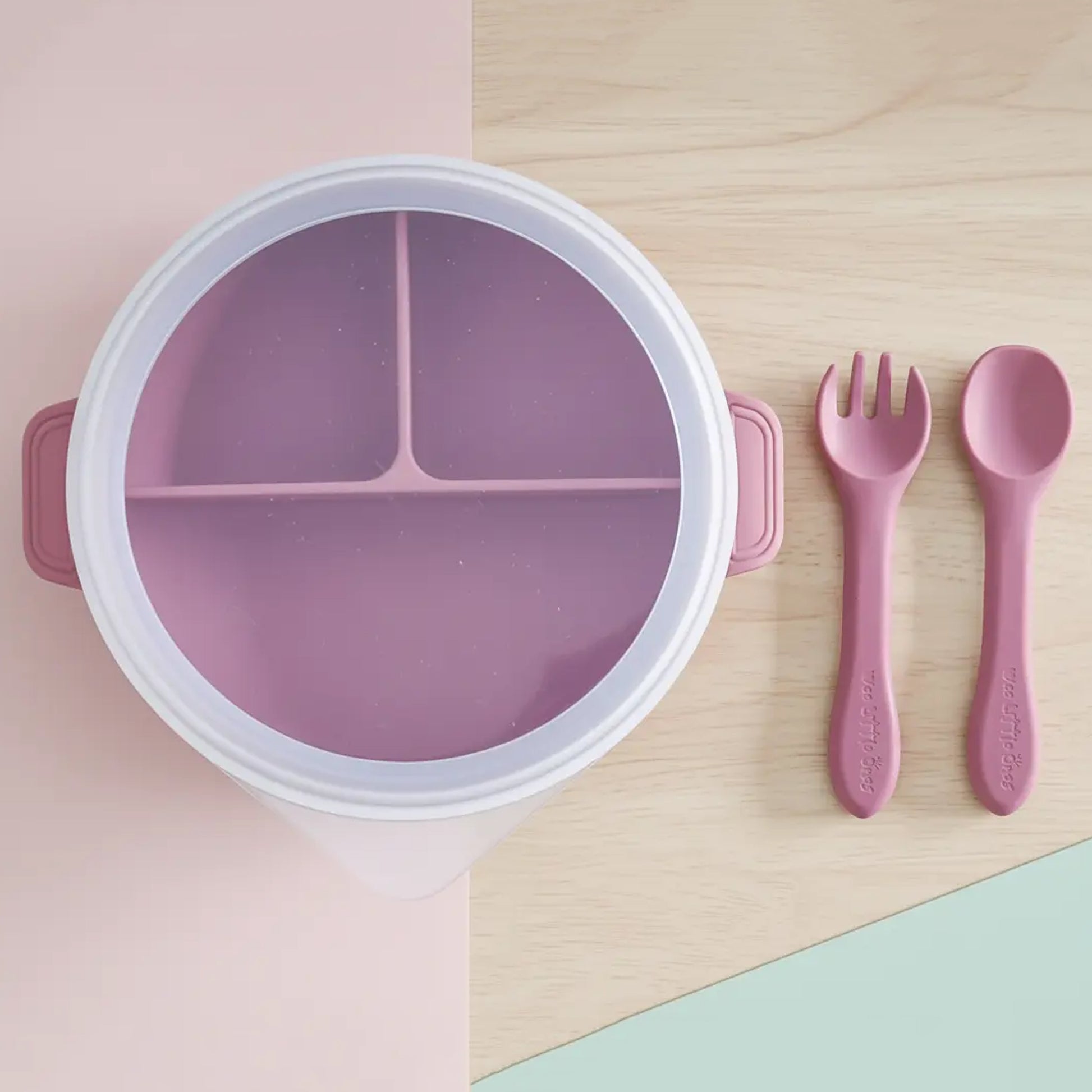 Round dark pink silicone divided plate with lid, matching spoon and fork set on wood and pastel background.