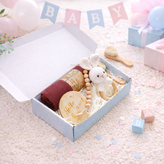 Baby gift set with toys and items in a box on a soft surface with 'BABY' banner in the background.
