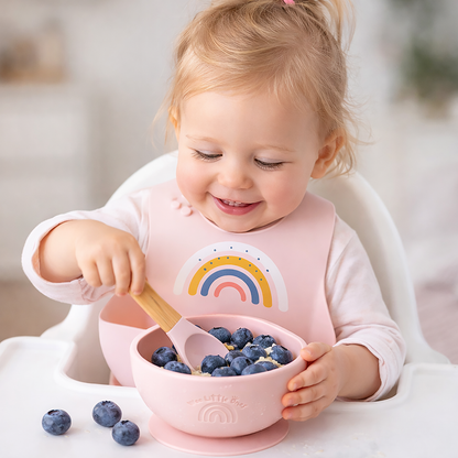 Child eating blueberries with a pink bowl and spoon, wearing a bib with a rainbow design.