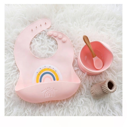 Pink baby bib with rainbow design, pink bowl with spoon, and textured pink object on white fluffy background