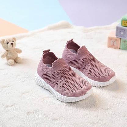 Pink breathable knit toddler shoes with soft white soles on a plush cream rug.