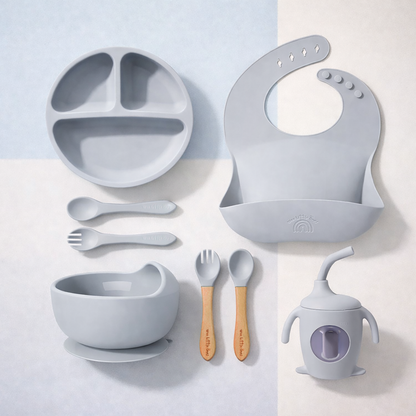 Set of gray baby feeding utensils including plate, bowl, spoons, and bib on a light background