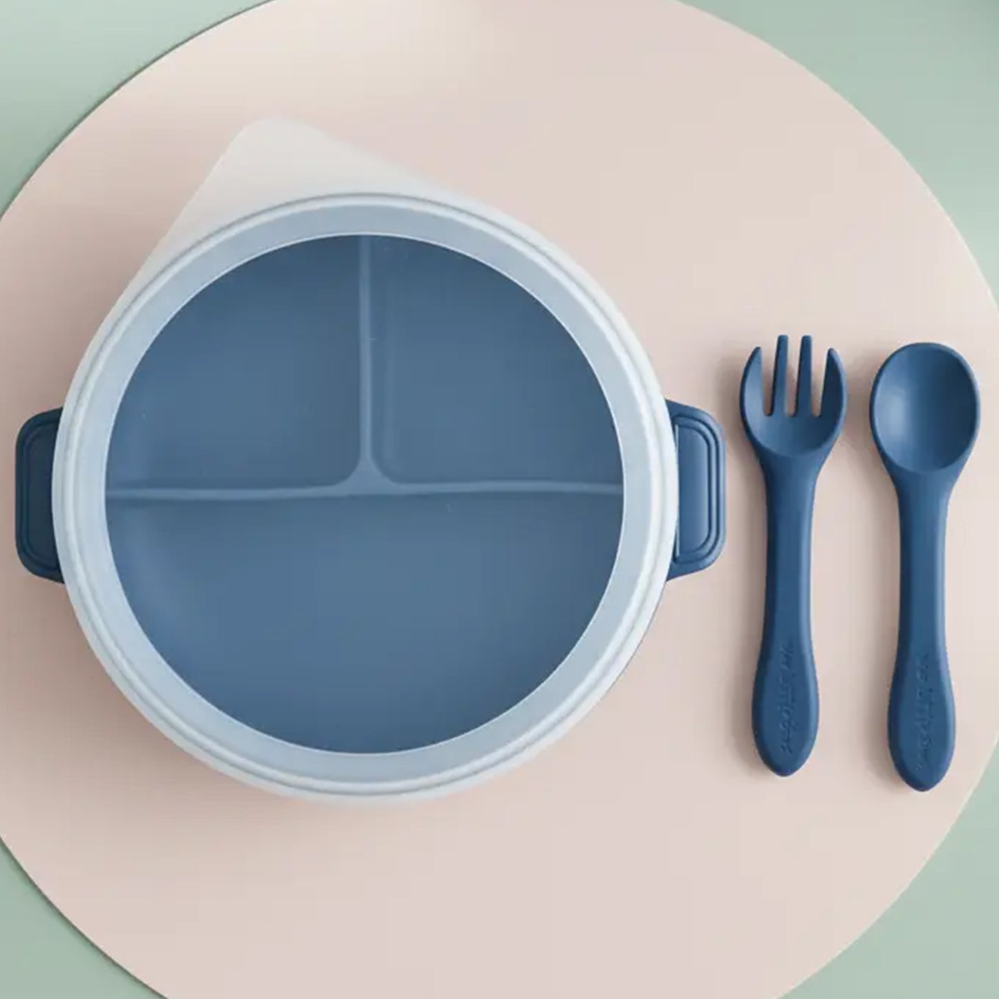 Navy blue silicone divided plate with handles beside matching fork and spoon on round beige and green surface.
