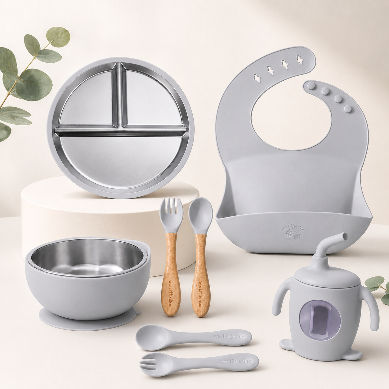 Collection of baby utensils and accessories including a bowl, spoons, fork, bib, and cup on a light background.