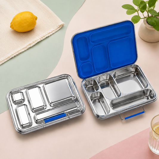 Two stainless steel compartment lunch boxes shown on a pastel background, one closed and one open to reveal a bright blue interior lid. A lemon, cloth, and small plant are styled around them.
