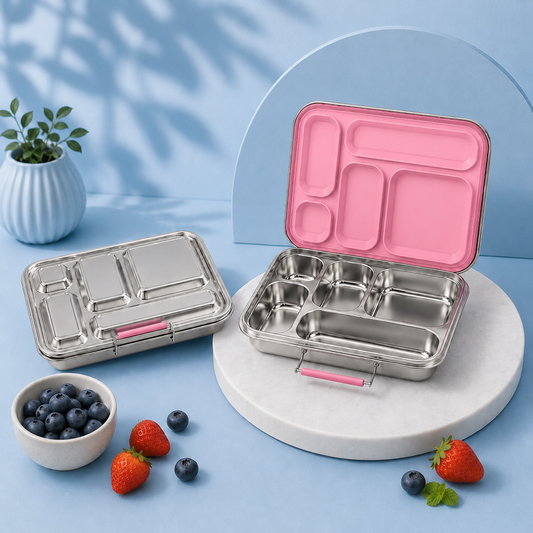 Two stainless steel compartment lunch boxes with pink lids are displayed on a light blue studio set. One box is closed and placed on the left, while the other is open and sits on a round pedestal on the right.