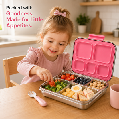 A young girl with a pink bow in her hair smiles while packing a stainless steel bento lunch box with a bright pink lid. The open box contains broccoli, tomatoes, blueberries, chickpeas, cheese cubes, and sandwich slices.