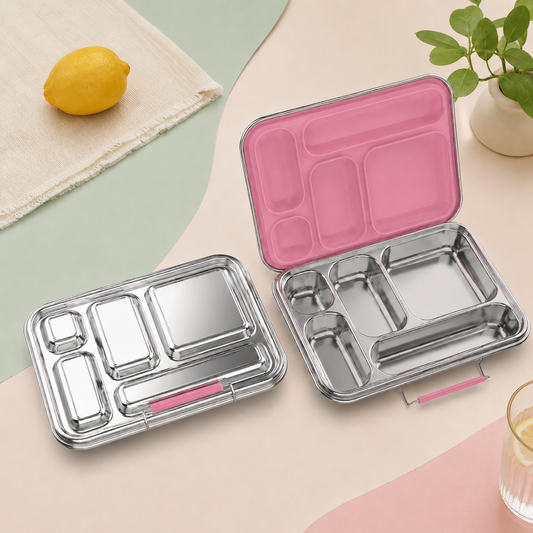 Two stainless steel bento lunch boxes with compartment dividers are shown on a pastel surface. One box is closed, revealing a pink interior lining and a small pink accessory slot, while the other is open to show the interior layout.
