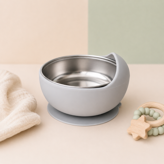 A baby feeding bowl with a matte gray silicone exterior, a stainless steel interior, and a suction base, styled on a beige surface with a cream textured cloth and a wooden star teether.