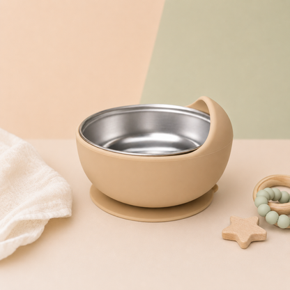 A beige baby feeding bowl with a stainless steel interior and suction base, styled on a pastel background with a white cloth and wooden teething toys.