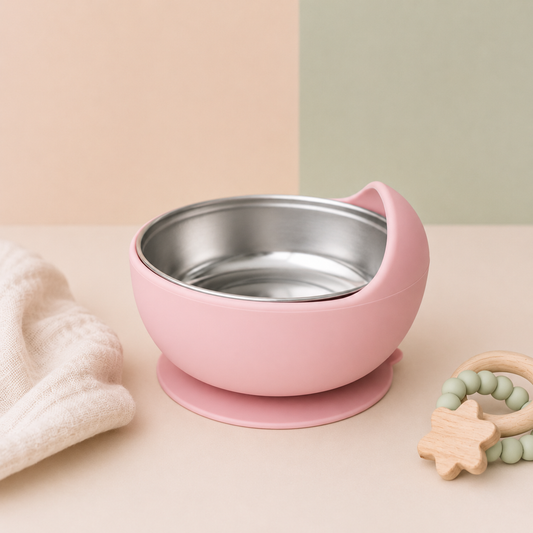 A pink silicone baby bowl with a stainless steel interior and suction base, styled on a beige surface with a soft cream cloth and a wooden bead teether in the background.