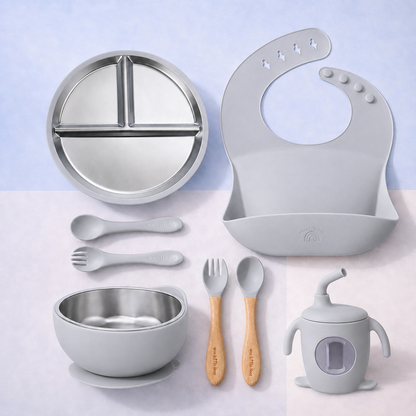 Collection of children's dining utensils and accessories including a plate, bowl, spoon, fork, bib, and cup on a light blue background.