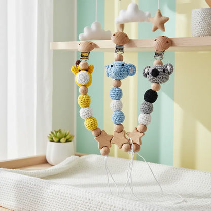 Crochet pacifier clips with yellow elephant, blue giraffe, and gray koala designs hanging on wooden rack.